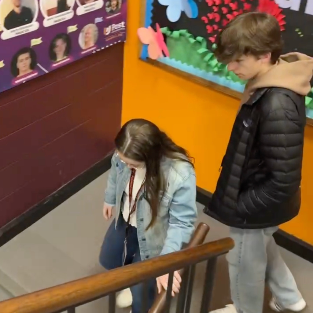 Penny and William are walking down a staircase indoors, with a bright orange wall and bulletin board behind them.