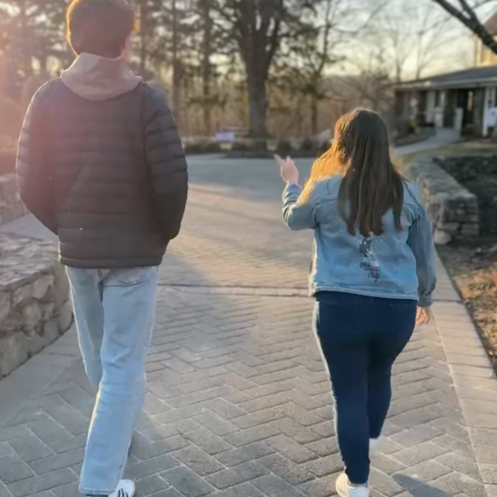 William and Penny walk outdoors on a paved path, with the sun setting behind them. Penny is gesturing as she speaks while Williams listens.