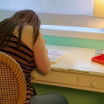 Penny sits at a wooden table in a softly lit room. She is writing in a notebook. A small table lamp with a white shade provides light. A red pencil case sits on the desk near the lamp.