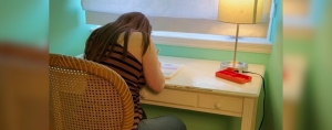 Penny sits at a wooden table in a softly lit room. She is writing in a notebook. A small table lamp with a white shade provides light. A red pencil case sits on the desk near the lamp.