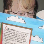 a cartoonized photo of Penny, with her glasses and light brown hair visible behind a book, is reading a colorful children's book.