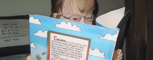 a cartoonized photo of Penny, with her glasses and light brown hair visible behind a book, is reading a colorful children's book.