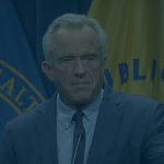 RFK Jr. stands at a podium with a microphone in front of flags from the U.S. Department of Health and Public Health Service.