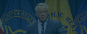 RFK Jr. stands at a podium with a microphone in front of flags from the U.S. Department of Health and Public Health Service.