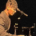 photo of Jon Batiste playing a grand piano on a dark stage