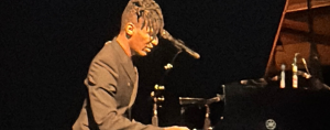photo of Jon Batiste playing a grand piano on a dark stage