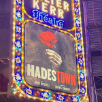 A brightly lit marquee for The Walter Kerr Theatre in New York City is shown at night. The sign features colorful, ornate lights framing a poster for the Broadway musical Hadestown. The poster depicts a hand holding a red flower against a dark, smoky background. The title “HADESTOWN” is prominently displayed