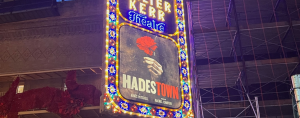 A brightly lit marquee for The Walter Kerr Theatre in New York City is shown at night. The sign features colorful, ornate lights framing a poster for the Broadway musical Hadestown. The poster depicts a hand holding a red flower against a dark, smoky background. The title “HADESTOWN” is prominently displayed