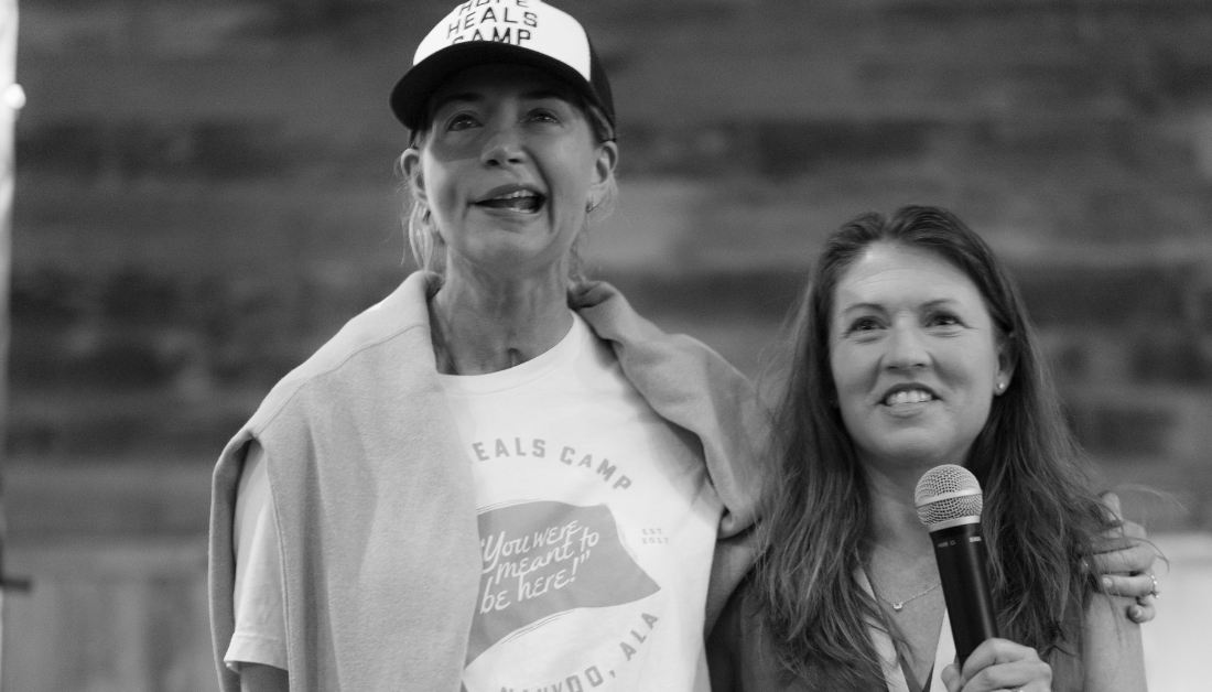 photo by Bryan Johnson of Amy Julia holding a mic and speaking as Katherine Wolf stands beside her and puts her arm around her