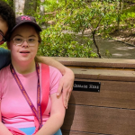 Penny sits on a wooden bench in a wooded park. She is smiling. Her boyfriend is sitting next to her, partially out of frame.