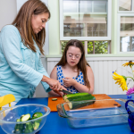 Penny and I are preparing food together in a kitchen. I'm showing Penny how to slice zucchini on a cutting board placed on a blue table. A glass baking dish and a bowl of chopped zucchini are nearby
