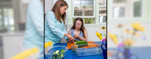 Penny and I are preparing food together in a kitchen. I'm showing Penny how to slice zucchini on a cutting board placed on a blue table. A glass baking dish and a bowl of chopped zucchini are nearby