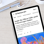 A tablet on a light wood desk displays a Washington Post article titled “Inside the Silicon Valley push to breed super-babies.” The subheading reads, “Investors say genetic prediction services for embryos, used by Elon Musk and others, are a trust fund for future children. Many scientists are skeptical.” The article is dated July 16, 2025, with an estimated 22-minute read time. The desk also features a pen, a white notebook, white earbuds, and a bunch of white and pink daisies in the corner.