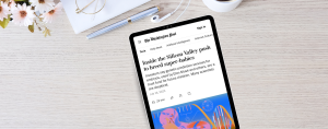 A tablet on a light wood desk displays a Washington Post article titled “Inside the Silicon Valley push to breed super-babies.” The subheading reads, “Investors say genetic prediction services for embryos, used by Elon Musk and others, are a trust fund for future children. Many scientists are skeptical.” The article is dated July 16, 2025, with an estimated 22-minute read time. The desk also features a pen, a white notebook, white earbuds, and a bunch of white and pink daisies in the corner.