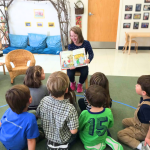 2015 photo of young Penny reading a book in a classroom to a group of children sitting in front of her