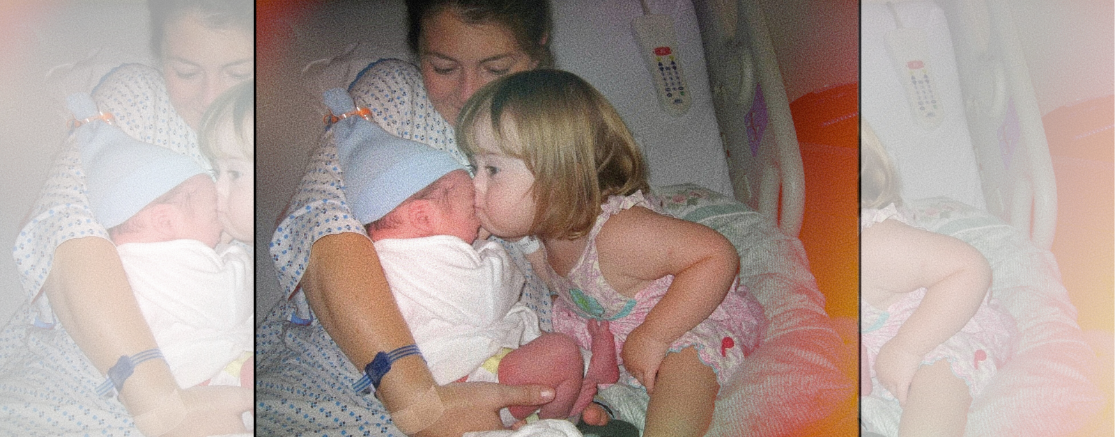 Amy Julia sits in a hospital bed wearing a hospital gown and holding a newborn baby wrapped in a white blanket and wearing a blue hat. Toddler Penny leans in to kiss the newborn on the lips while the Amy Julia looks on with a gentle smile.