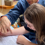 2023 photo of Peter sitting next to Penny. They are at a table covered with papers, pens, and a coffee cup. He’s pointing to a document as she is focused on reading it