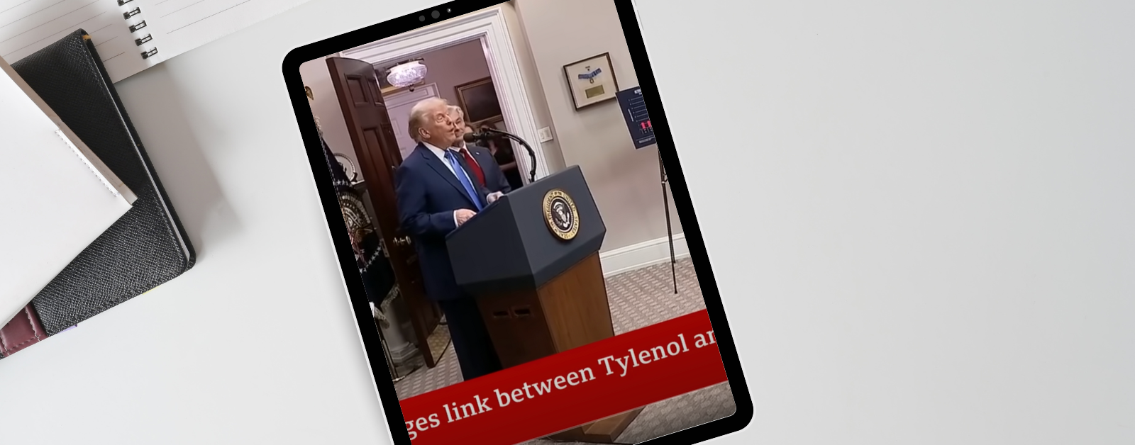Tablet on a desk displays a video of former President Donald Trump speaking at a podium with the presidential seal. Another person stands behind him. A red news ticker partially visible at the bottom reads '…ges link between Tylenol an…'. Notebooks and folders are arranged on the desk next to the tablet.