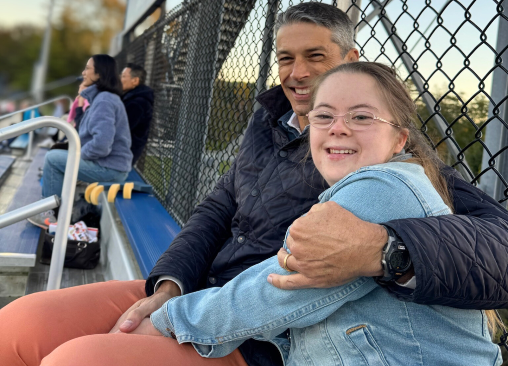 Peter and Penny sit together on metal bleachers, smiling warmly at the camera. Peter has his arm wrapped around Penny. They are both wearing jackets. A chain-link fence and other spectators are visible in the background. Peter and Penny sit on bleachers keeping each other warm during soccer game