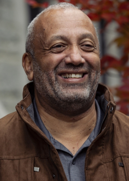 A smiling John Swinton, who is a disability theologian, stands outdoors wearing a brown jacket, with red autumn leaves and a stone wall in the background