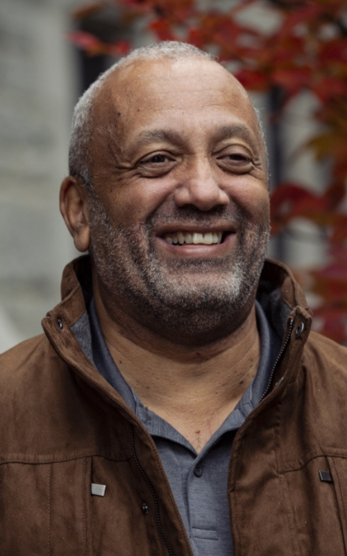 A smiling John Swinton, who is a disability theologian, stands outdoors wearing a brown jacket, with red autumn leaves and a stone wall in the background
