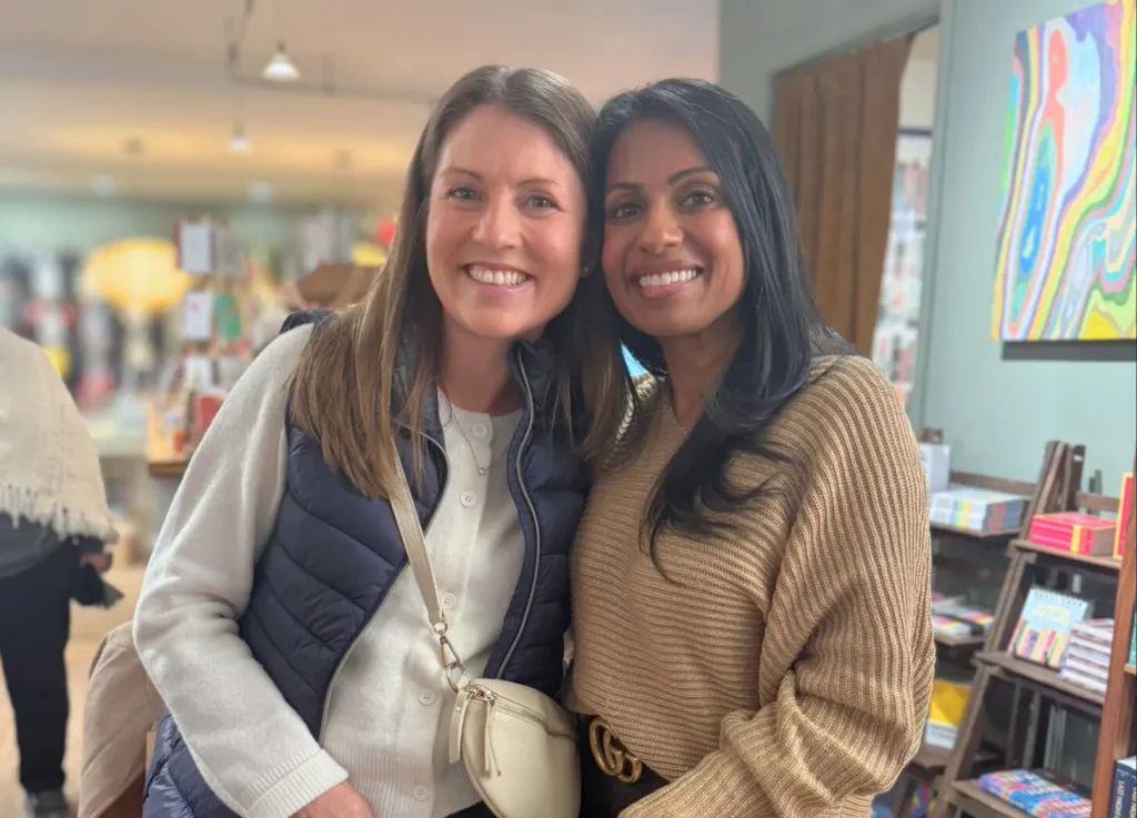 Amy Julia and Niro stand close together in a cozy bookshop, smiling at the camera. Colorful artwork and shelves of books are in the background.