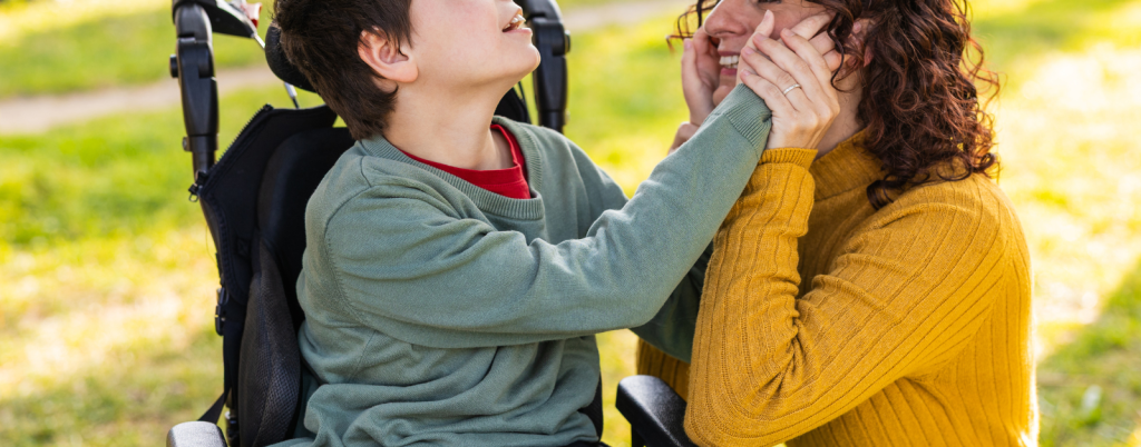 child playing with mother outdoors. The child is using a wheelchair, and they both are using their hands to see the others face.