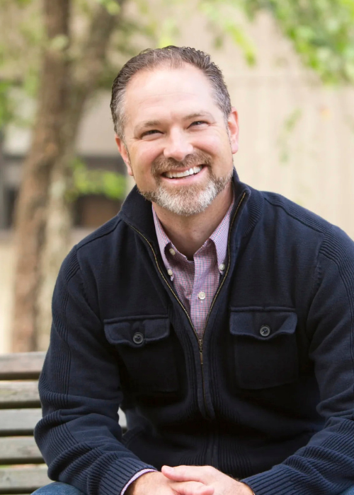 A smiling Kelly Kapic sits outdoors on a bench