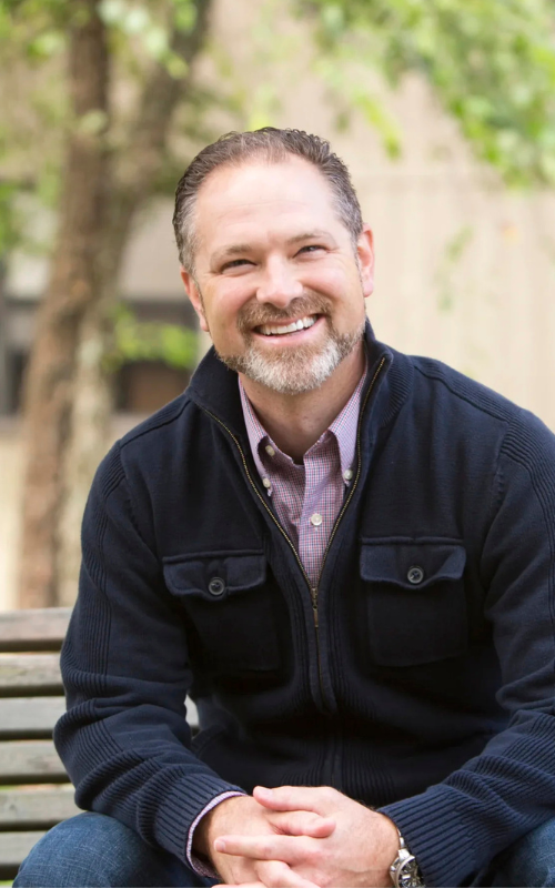 A smiling Kelly Kapic sits outdoors on a bench