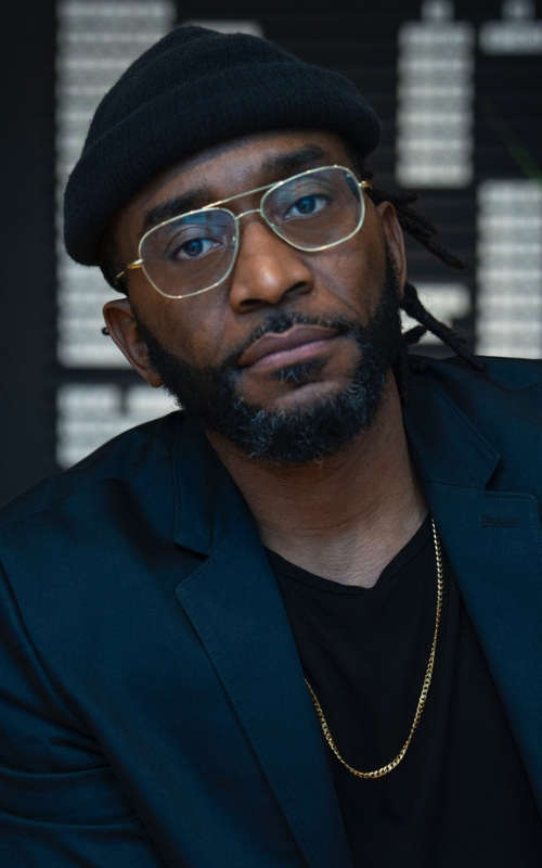 a portrait of Sho Baraka with book shelves behind him