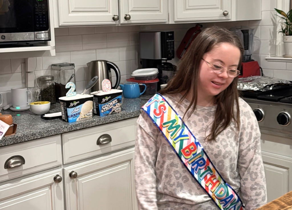 Penny stands in the kitchen wearing a birthday sash. Behind her are two candles affixed to the makings for ice cream sundaes. The candles are a two and a zero. Penny stands in the kitchen wearing a birthday sash. Behind her are two candles affixed to the makings for ice cream sundaes. The candles are a two and a zero.