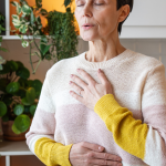 Joyful calm mature woman meditating with deep breathing, hands rest on chest. Breathing for relax