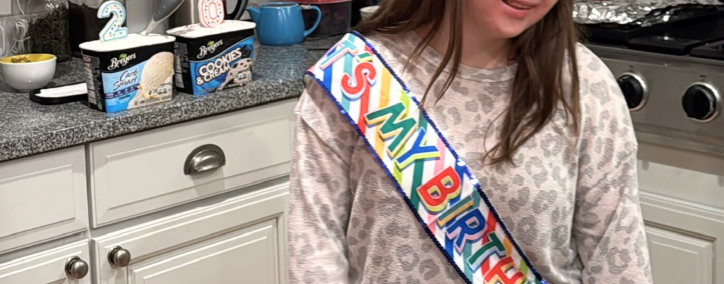 Penny stands in the kitchen wearing a birthday sash. Behind her are two candles affixed to the makings for ice cream sundaes. The candles are a two and a zero.