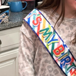 Penny stands in the kitchen wearing a birthday sash. Behind her are two candles affixed to the makings for ice cream sundaes. The candles are a two and a zero.