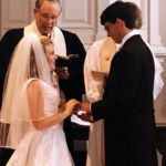 Peter and I at the front of a church during our wedding ceremony, standing before an officiant at the altar, with attendants beside us and guests seated in pews decorated with white bows and flowers.