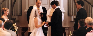 Peter and I at the front of a church during our wedding ceremony, standing before an officiant at the altar, with attendants beside us and guests seated in pews decorated with white bows and flowers.