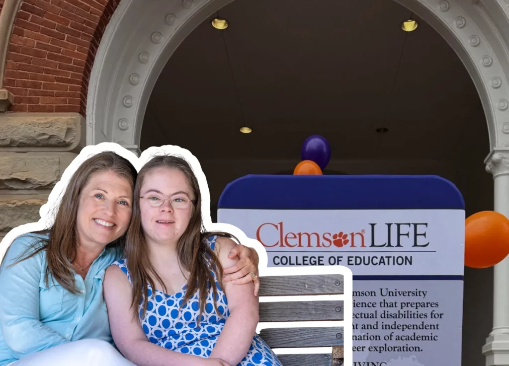 Amy Julia sits close to Penny with her arm around her on a bench in front of a ClemsonLIFE sign at Clemson University. The pair are framed by a large brick archway, with purple and orange balloons in the background. Amy Julia sits close to Penny with her arm around her on a bench in front of a ClemsonLIFE sign at Clemson University. The pair are framed by a large brick archway, with purple and orange balloons in the background.