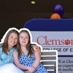 Amy Julia sits close to Penny with her arm around her on a bench in front of a ClemsonLIFE sign at Clemson University. The pair are framed by a large brick archway, with purple and orange balloons in the background.
