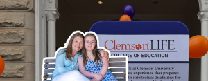Amy Julia sits close to Penny with her arm around her on a bench in front of a ClemsonLIFE sign at Clemson University. The pair are framed by a large brick archway, with purple and orange balloons in the background.