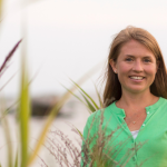 I smile at the camera while standing outdoors among tall green grasses, wearing a light green cardigan and a white top, with a soft, blurred marsh behind me