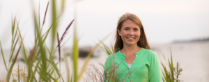I smile at the camera while standing outdoors among tall green grasses, wearing a light green cardigan and a white top, with a soft, blurred marsh behind me