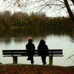 2 people sitting on a bench near a pond