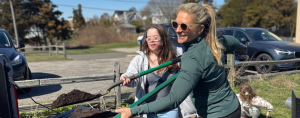 Penny and her aunt work together outdoors, shoveling soil out of the back of a truck on a sunny day.