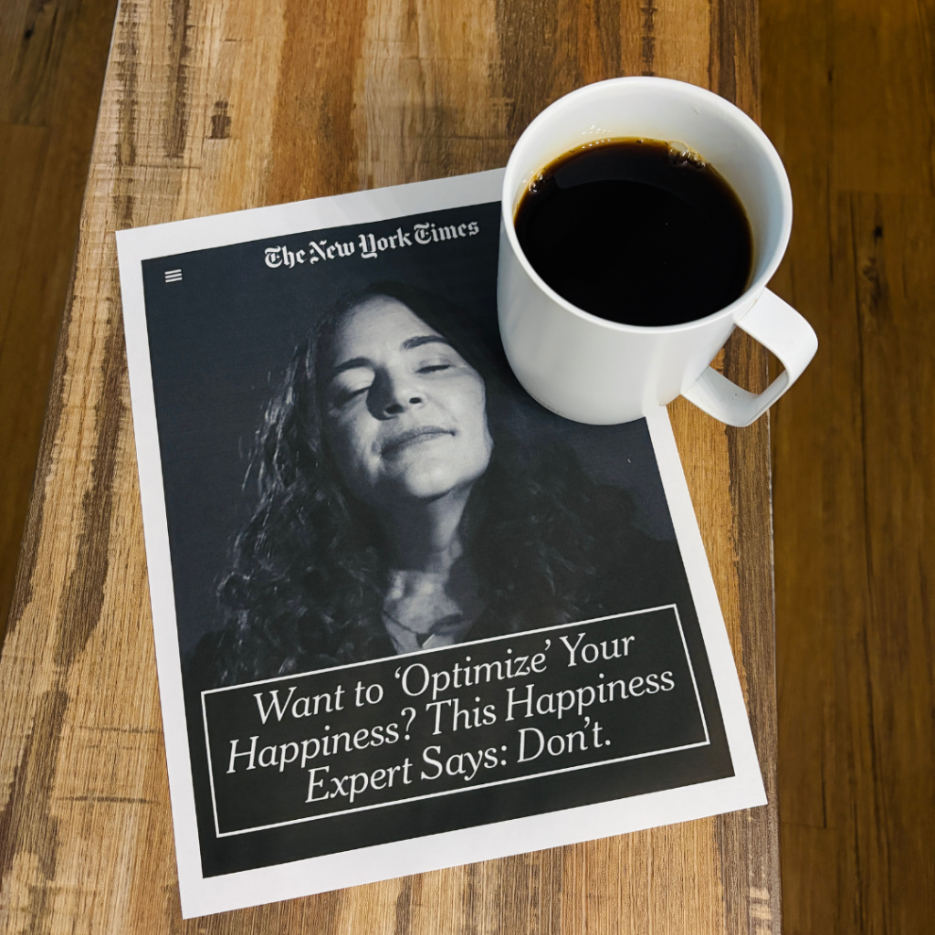 A white mug of black coffee sits on a wooden table beside a printed New York Times article. The article features a black-and-white portrait and the headline, “Want to ‘Optimize’ Your Happiness? This Happiness Expert Says: Don’t.”