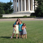 2015 photo of Amy Julia wearing sunglasses and standing on a grassy lawn in front of the Jefferson Memorial. She is smiling and hugging three Penny, William, and Marilee, who are standing close together and facing the camera on a sunny day with a clear blue sky