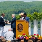 William receives his diploma on stage. Hills and a lake are behind him, and seated graduates are in the foreground