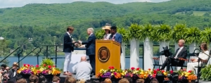 William receives his diploma on stage. Hills and a lake are behind him, and seated graduates are in the foreground