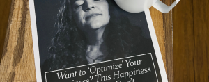 A white mug of black coffee sits on a wooden table beside a printed New York Times article. The article features a black-and-white portrait and the headline, “Want to ‘Optimize’ Your Happiness? This Happiness Expert Says: Don’t.”