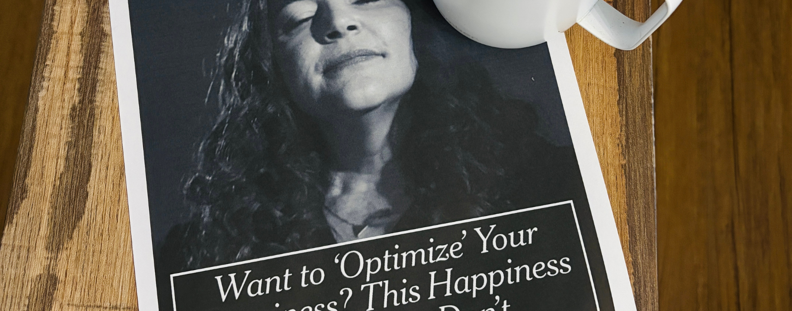 A white mug of black coffee sits on a wooden table beside a printed New York Times article. The article features a black-and-white portrait and the headline, “Want to ‘Optimize’ Your Happiness? This Happiness Expert Says: Don’t.”