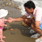 Peter crouches at the edge of the ocean, smiling and reaching out to toddler Penny standing on a beach. Waves wash around the his feet while Penny, wearing a pink patterned swimsuit and white sun hat, faces him near the shoreline.